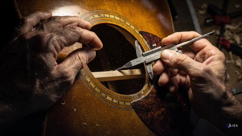 Master violin maker's hands examining the internal bracing structure of an acoustic guitar through the sound hole.